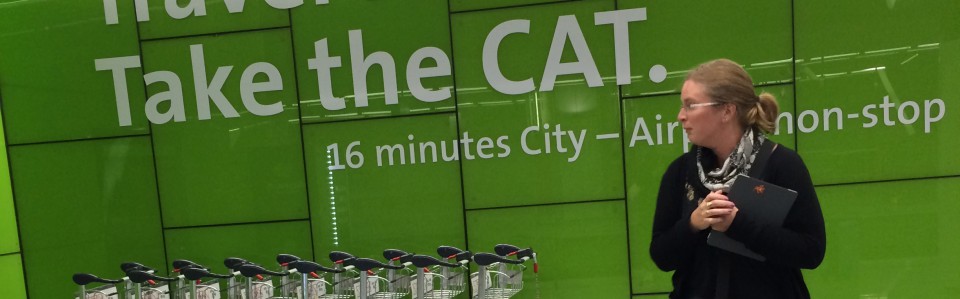 Image of woman in foreground, airport trollies and bright background. Text on image says Take the CAT, 16 minutes City to Airport non-stop. Describes train service in Vienna from city to airport,